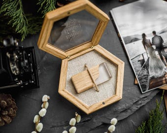Puede incluir: Una caja hexagonal de madera con tapa de cristal que presenta los nombres Daniel y Emma grabados. Dentro, una memoria USB rectangular de madera y resina transparente descansa sobre un forro de tela beige, sujeta por una barra de madera.