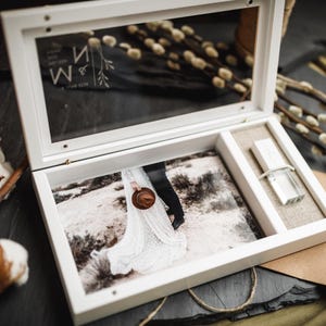 May include: A white wooden keepsake box with a glass lid featuring the engraved letters M and N. The interior contains a printed photograph of a person in a white dress holding a hat, alongside a small rectangular USB flash drive.