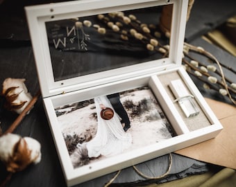 Puede incluir: Una caja de recuerdos de madera blanca con tapa de cristal grabada con las letras M y N. El interior contiene una fotografía impresa de una persona con vestido blanco sosteniendo un sombrero, junto a una pequeña memoria USB rectangular.