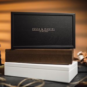 May include: Three rectangular boxes in black, brown, and white are stacked. The top box is black and features the names "JULIA & DANIEL" and the date "26.06.2026" in gold lettering. A vintage camera is on the left.