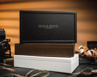 Puede incluir: Una pila de tres cajas rectangulares en negro, marrón y blanco. La caja superior es negra y tiene los nombres "JULIA & DANIEL" y la fecha "26.06.2026" en letras doradas. Se ve una cámara vintage.
