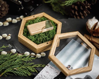 Puede incluir: Una memoria USB de madera en una caja hexagonal de madera llena de musgo verde. La caja está abierta, revelando la memoria USB. Otro marco hexagonal de madera vacío está al lado, con una foto dentro. La escena está decorada con vegetación y una flor de algodón.
