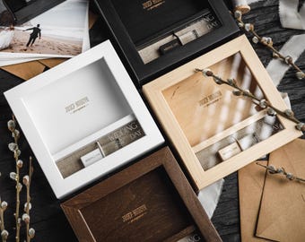 Puede incluir: Una colección de marcos de fotos cuadrados de madera en varios colores: blanco, negro, marrón claro y marrón oscuro. Cada marco tiene un área empotrada con una tela tipo lino y pequeñas etiquetas de madera. Los marcos están dispuestos sobre una superficie de madera oscura.