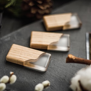 Può includere: Tre chiavette USB in legno con intarsio in resina trasparente sono disposte su una superficie grigio scuro. Le chiavette USB hanno una finitura a venatura naturale. Lo sfondo include una fotocamera, rami di salice e una pigna.