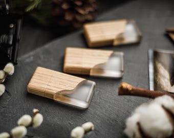 Puede incluir: Tres unidades flash USB de madera con una incrustación de resina transparente están dispuestas sobre una superficie gris oscuro. Las unidades flash tienen un acabado de veta de madera natural. El fondo incluye una cámara, ramas de sauce y una piña.
