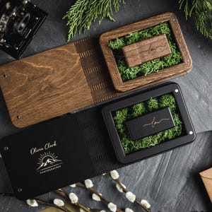 Könnte beinhalten: Hölzerne und schwarze USB-Sticks mit dem eingravierten Namen "law", präsentiert in dekorativen Boxen mit grünem Moos. Die Boxen werden mit einem Fotoalbum aus Holz und einem schwarzen Fotoalbum präsentiert, auf denen der Name "Olivia Clark Photographer" aufgedruckt ist.