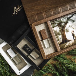 Peut inclure: Un coffret photo en bois avec une photographie de mariage, un album photo noir avec des lettres dorées et deux clés USB. Le coffret photo a un couvercle à charnière et un intérieur en lin. Les clés USB sont dans des étuis blancs et noirs.