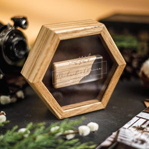 May include: A wooden hexagonal display case with a clear front showcases a rectangular object with the inscription "Michael & Olivia". The case is set against a dark background with a vintage camera and decorative elements.