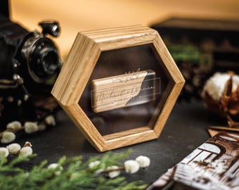 Puede incluir: Un expositor hexagonal de madera con frente transparente exhibe un objeto rectangular con la inscripción "Michael & Olivia". El estuche se encuentra sobre un fondo oscuro con una cámara vintage y elementos decorativos.