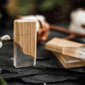 May include: A rectangular lighter with a wooden top and a clear, transparent bottom. The lighter is made of wood and clear plastic. The lighter is sitting on a dark, textured surface. There are other lighters in the background.