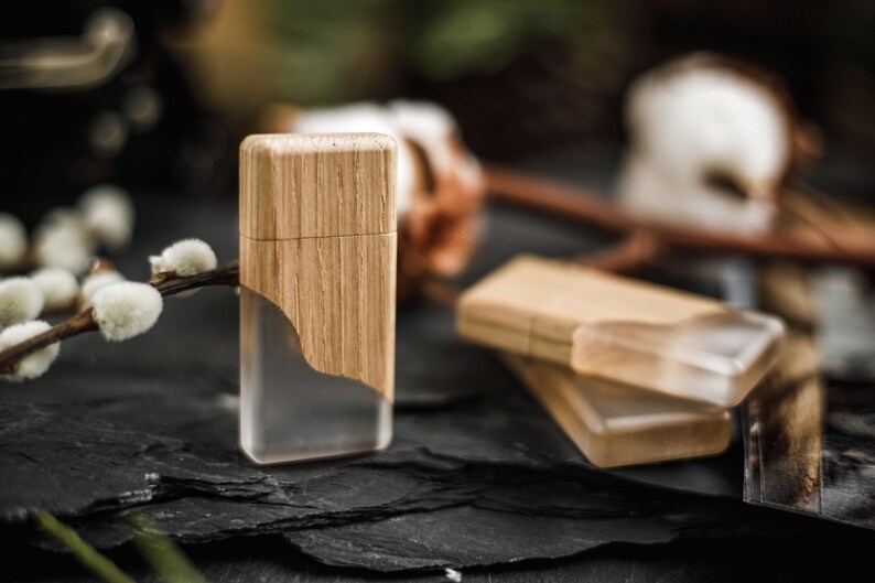 May include: A rectangular lighter with a wooden top and a clear, transparent bottom. The lighter is made of wood and clear plastic. The lighter is sitting on a dark, textured surface. There are other lighters in the background.
