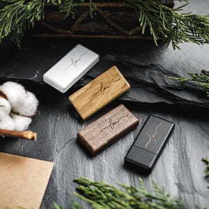 Könnte beinhalten: Vier USB-Sticks in Weiß, hellem Holz, dunklem Holz und Schwarz. Jeder Stick trägt das Wort "love" in kursiver Schrift. Die Sticks sind auf einer dunklen, strukturierten Oberfläche mit Zweigen und einer Baumwollkapsel angeordnet.