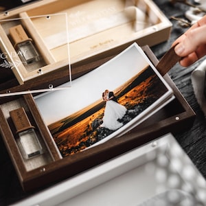 Può includere: Scatole di foto in legno con coperchio in acrilico trasparente e chiavetta USB. Le scatole contengono foto di matrimonio. L'immagine mostra una persona che apre la scatola per rivelare le foto. Le scatole sono su una superficie di legno scuro.