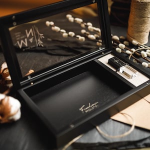 Pode incluir: Uma caixa de madeira preta com tampa de vidro articulada com as letras M e N e um design floral. O interior é preto com um pequeno compartimento retangular contendo uma unidade flash USB preta e transparente e texto dourado.