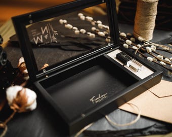 Puede incluir: Una caja de madera negra con tapa de cristal abatible que muestra las letras M y N con un diseño floral. El interior es negro con un pequeño compartimento rectangular que contiene una memoria USB negra y transparente y texto dorado.