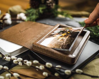 Puede incluir: Un álbum de fotos de madera con una cubierta de acrílico transparente, que muestra una fotografía. El álbum está abierto, revelando la imagen, y está rodeado de otros álbumes y elementos decorativos. El álbum es de madera.