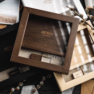 Può includere: Tre scatole di legno con piani in vetro, in nero, marrone e tonalità legno chiaro. Le scatole hanno un design a cornice quadrata e contengono un interno in lino. Il testo "WEDDING DAY" è visibile su un'etichetta. Scatole per fotografia.