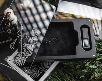 Puede incluir: Una caja de madera negra con un pequeño frasco de perfume plateado en su interior. Tarjetas de acrílico transparente con diseños botánicos blancos y texto están superpuestas en la parte delantera. El texto en la tarjeta dice "Patricia & Christian 21/06/2026".