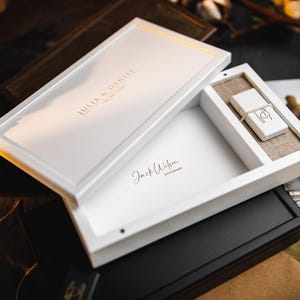 Könnte beinhalten: Weiße Holz-Präsentationsbox, teilweise geöffnet, mit einem USB-Stick und einem Fach. Auf der Box stehen die Namen "JULIA & DANIEL" und das Datum "25.06.2022". Die Signatur des Fotografen "Jack Wilson photography" ist ebenfalls sichtbar.