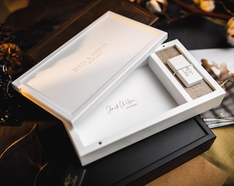 Puede incluir: Caja de presentación de madera blanca, parcialmente abierta, que revela una unidad USB y un compartimento. La caja está inscrita con los nombres "JULIA & DANIEL" y la fecha "25.06.2022". También es visible la firma del fotógrafo "Jack Wilson photography".