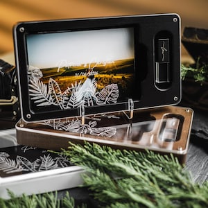 Pode incluir: Uma caixa de exibição de fotos preta e de madeira com uma foto de paisagem e um design de folhas gravadas. Uma câmera vintage e um pequeno baú de madeira estão no fundo. A caixa tem um slot para uma unidade USB.