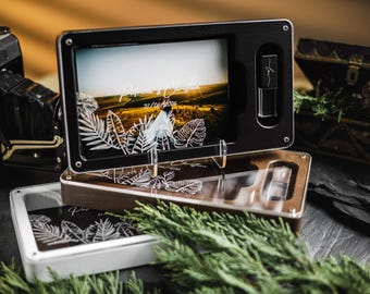 Puede incluir: Una caja de exhibición de fotos negra y de madera con una foto de paisaje y un diseño de hojas grabadas. Una cámara vintage y un pequeño cofre de madera están en el fondo. La caja tiene una ranura para una unidad USB.