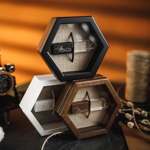 May include: Three hexagonal display boxes in black, white, and brown, stacked. Each box contains a small, rectangular object with script text. A vintage camera and cotton flower are in the foreground.