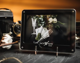Puede incluir: Un marco de fotos negro con temática de boda. La foto presenta copas de champán, un ramo de flores blancas y tarjetas. El marco tiene los nombres "Jack Rose" y la fecha 28/11/2016. Una cámara vintage y tallos de algodón están en el fondo.