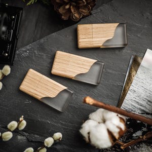 May include: Three wooden USB flash drives with a clear, curved resin inlay. The drives are arranged on a dark grey surface with decorative elements including cotton bolls, a vintage camera, and a pine cone. The wood grain is visible.
