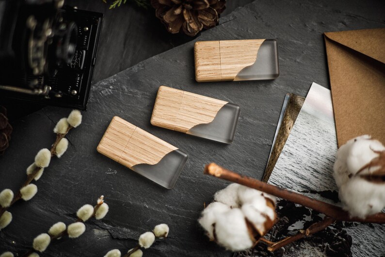 May include: Three wooden USB flash drives with a clear, curved resin inlay. The drives are arranged on a dark grey surface with decorative elements including cotton bolls, a vintage camera, and a pine cone. The wood grain is visible.