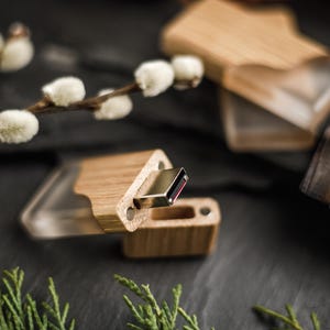 May include: A wooden USB flash drive with a clear plastic cover. The drive is made of light-coloured wood and has a metal connector. The image also shows a wooden box and sprigs of greenery.