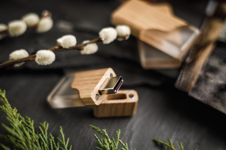May include: A wooden USB flash drive with a clear plastic cover. The drive is made of light-coloured wood and has a metal connector. The image also shows a wooden box and sprigs of greenery.