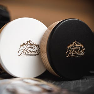 May include: Two circular wooden containers, one white and one black, with the text "James Mitchell Photographer" in a mountain-themed logo. The containers have a wooden rim and a flat top. The background is blurred.