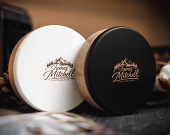 Puede incluir: Dos recipientes redondos de madera, uno blanco y otro negro, con el texto "James Mitchell Photographer" en un logotipo con temática de montaña. Los recipientes tienen un borde de madera y una parte superior plana. El fondo está borroso.