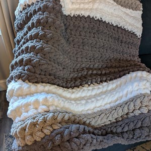 May include: A chunky knit blanket in shades of gray, white, and tan. The blanket is draped over a dark gray surface, showcasing its textured, oversized stitches. The color scheme creates a cozy, neutral aesthetic, suitable for home decor.