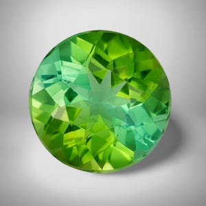 May include: A round, faceted gemstone with a vibrant green hue. The gem has a star-like pattern in the center and is cut with multiple facets, creating a sparkling effect. The gemstone is set against a white background.