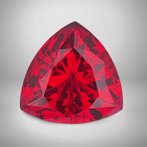 May include: A vibrant, triangular-cut gemstone in a rich red colour. The surface is meticulously faceted, producing a multitude of reflective surfaces that capture and scatter light. The gem is precisely shaped as an equilateral triangle, highlighting its clarity and sparkle.