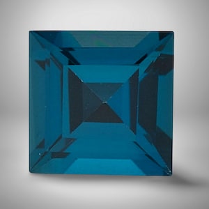 Puede incluir: Una gema de color turquesa, tallada en forma cuadrada, con un diseño facetado. La gema tiene un centro oscuro, casi negro, y una superficie pulida y reflectante. La gema se encuentra sobre un fondo gris degradado.