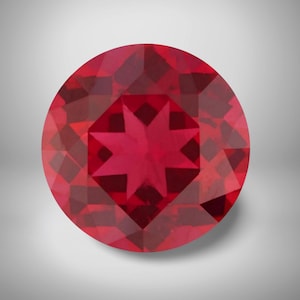 May include: A round, faceted gemstone with a deep red hue. The gem features a star-shaped pattern in the center, surrounded by geometric facets. The gemstone is set against a soft, neutral background.
