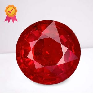 Könnte beinhalten: Ein facettierter, rund geschliffener, tiefroter Edelstein. Der Edelstein hat eine brillante, reflektierende Oberfläche mit mehreren dreieckigen Facetten. Ein goldenes Siegel mit einem Häkchen und rosa Bändern befindet sich in der oberen linken Ecke.
