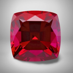 May include: A faceted, cushion-cut gemstone with a deep red hue. The gem's surface reflects light, showcasing its intricate internal structure. The square shape has rounded corners, and the gem is set against a neutral background.