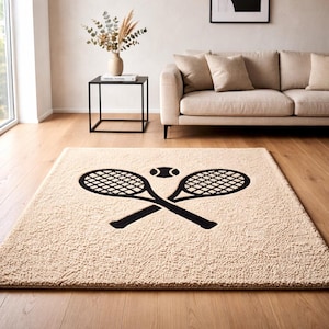 Puede incluir: Una alfombra rectangular beige con un diseño de raqueta y pelota de tenis negra. Dos raquetas de tenis cruzadas y una pelota de tenis están centradas en la alfombra. La alfombra está colocada sobre un suelo de madera en un salón.