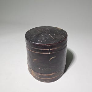 May include: A cylindrical dark brown wooden box with a removable lid. The surface features visible signs of wear, including light scratches and scuffs, with subtle horizontal banding details around the body and the top edge of the lid.