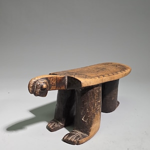Könnte beinhalten: Ein niedriger Holzhocker mit einem geschnitzten Tierkopf an einem Ende und vier stabilen Beinen. Die Sitzfläche ist oval geformt und weist an den Rändern eingravierte geometrische Muster auf. Das Holz hat ein dunkelbraunes Finish an den Beinen und am Kopf, während die Sitzfläche einen helleren Naturholzton aufweist. Die Konstruktion ist massiv und handgeschnitzt mit sichtbarer Textur.
