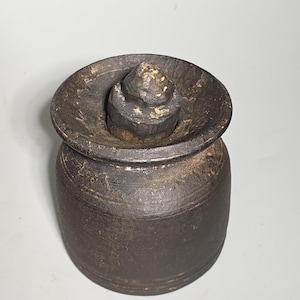 May include: A dark brown, antique-style butter churn with a lid. The churn is cylindrical with a slightly flared rim and a wooden knob on top. The wood has a weathered appearance.