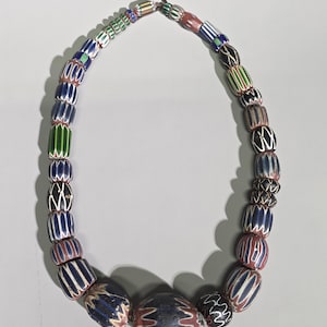 May include: A necklace composed of various barrel-shaped glass beads featuring intricate chevron and striped patterns in colours including navy blue, white, green, black, and terracotta red, strung together on a thin cord with a knotted closure.