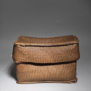 May include: A rectangular, woven basket with a hinged lid. The basket is made of brown, natural fibers, and the weave creates a textured surface. The lid is slightly curved, and the basket appears to be empty. The basket is a storage container.