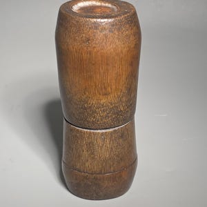 Puede incluir: Un recipiente de madera cilíndrico de dos piezas con un acabado marrón cálido natural. La sección superior es ligeramente más pequeña que la inferior, y la veta de la madera es visible. El recipiente parece hecho a mano.