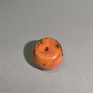 Könnte beinhalten: Eine runde, orangefarbene Perle mit glatter, polierter Oberfläche und einem zentralen Loch. Die Perle weist unregelmäßige dunkelbraune und schwarze Einschlüsse auf, die über die Oberfläche verteilt sind und ein natürliches, gesprenkeltes Aussehen mit glänzendem Finish erzeugen.