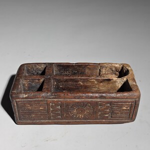 May include: A rectangular dark brown wooden organizer box with five interior compartments of varying sizes. The front exterior panel features hand-carved geometric patterns and a central circular sunburst motif with a weathered texture.